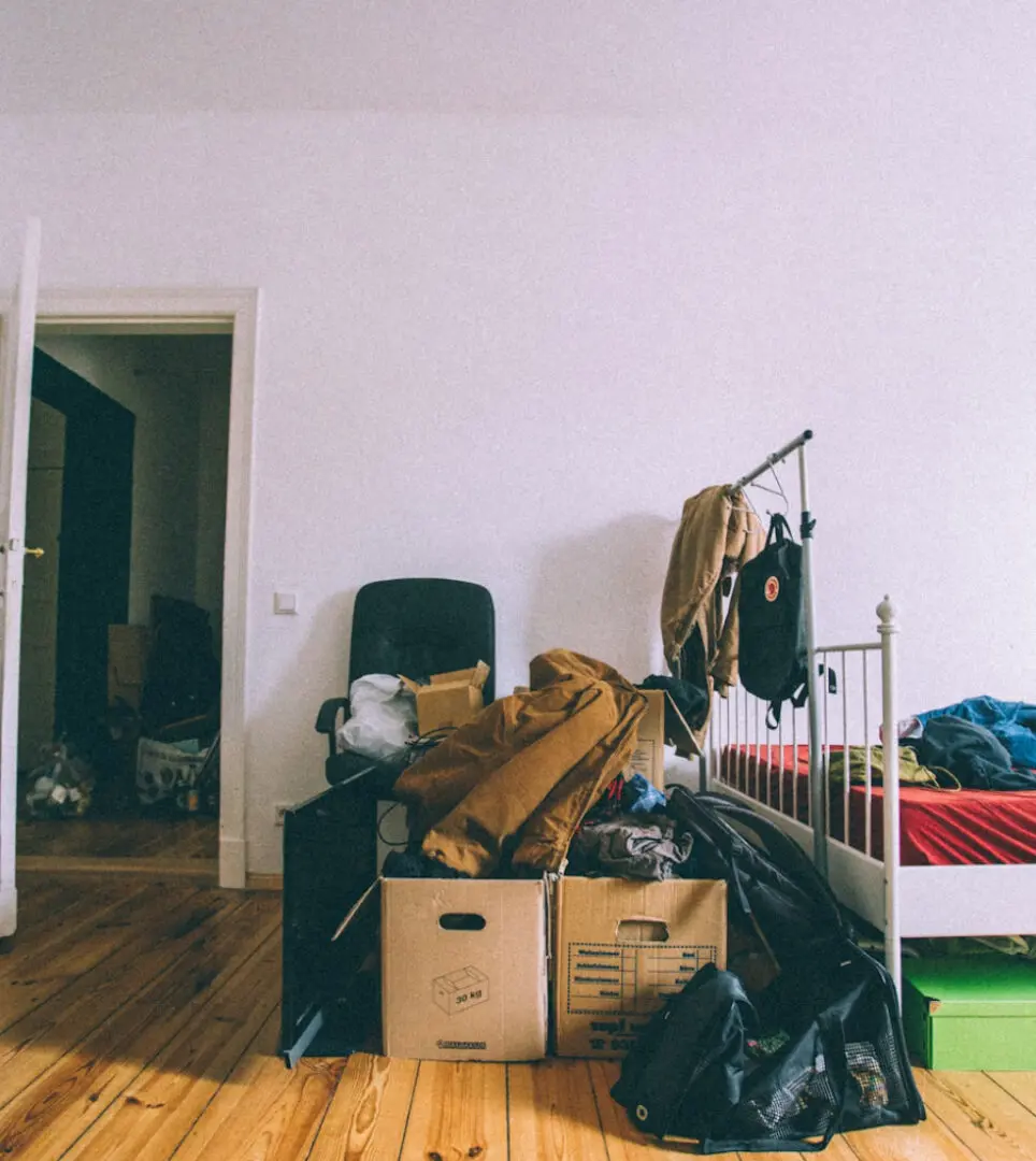 A cluttered room with boxes, clothes, and furniture against a white wall.