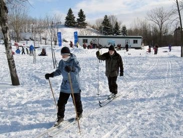 Two people skiing on a snowy trail with trees and buildings in the background.
