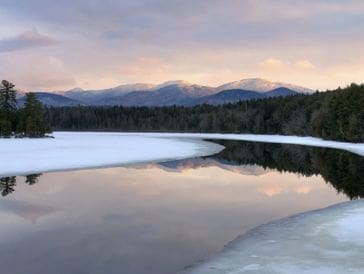 Tranquil partially frozen lake reflecting mountains and sky at dusk.
