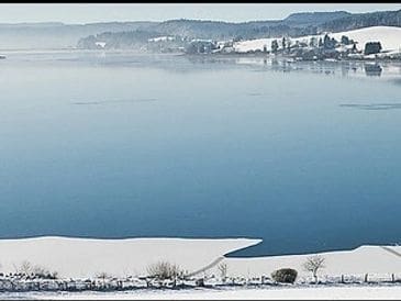 A snowy lakeside landscape with calm blue water and distant hills.