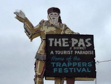 A wooden statue of a figure holding a sign for The Pas Trappers Festival.
