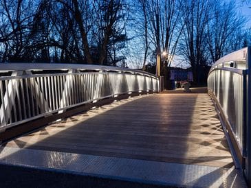 A well-lit pedestrian bridge at dusk with bare trees in the background.