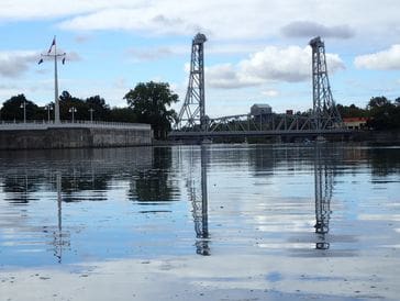 Calm river reflecting industrial towers and buildings under a partly cloudy sky.