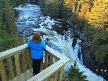 Person in blue jacket overlooking a rushing river from a wooden platform.