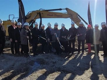 Group of people in hard hats breaking ground with shovels in front of excavator.