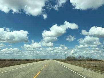 Empty road stretches under a blue sky with fluffy clouds.