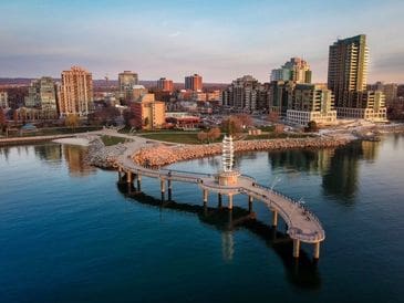 Curved pier with lighthouse extending into calm water, city skyline in background at sunset.
