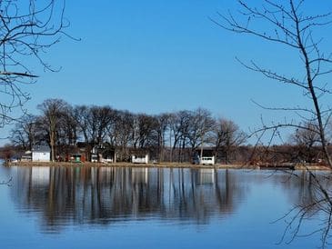 Calm lake reflecting trees and houses under a clear blue sky.