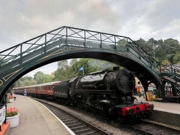 A vintage steam train passing under a metal footbridge at a station.