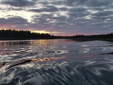 Sunset over a calm lake with rippling water and forest silhouette.