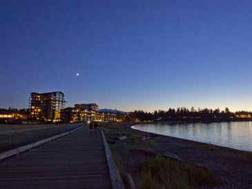 Evening view of a boardwalk beside water with city lights and a crescent moon.