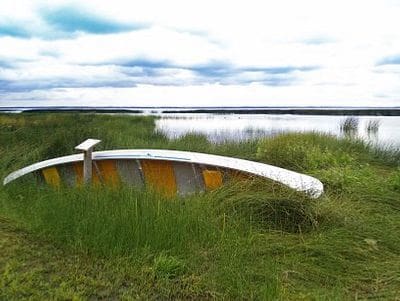 An overturned white rowboat resting in tall grass near a calm body of water under a cloudy sky.