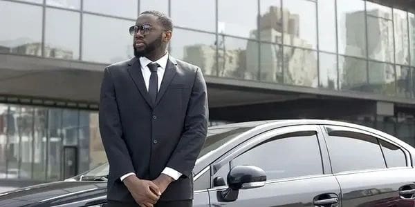 A man in a black suit and sunglasses stands confidently by a black car.