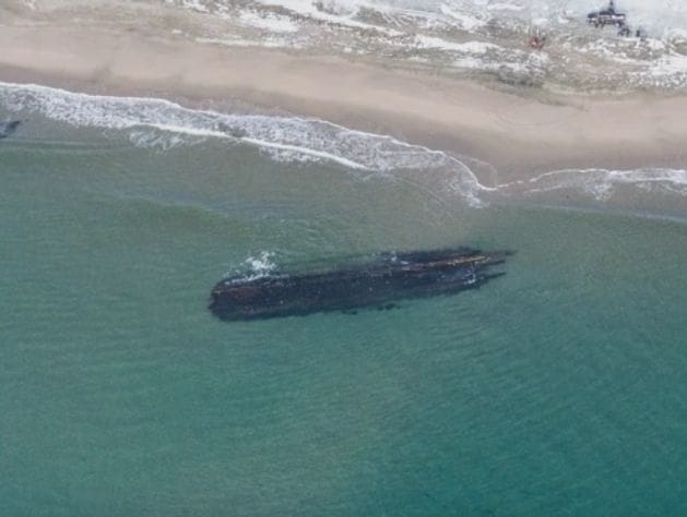 A shipwreck stranded near a sandy beach in shallow waters.