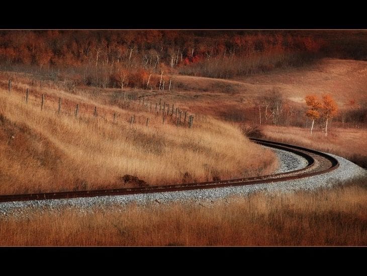 A winding road curves through autumn-colored fields under a misty sky.