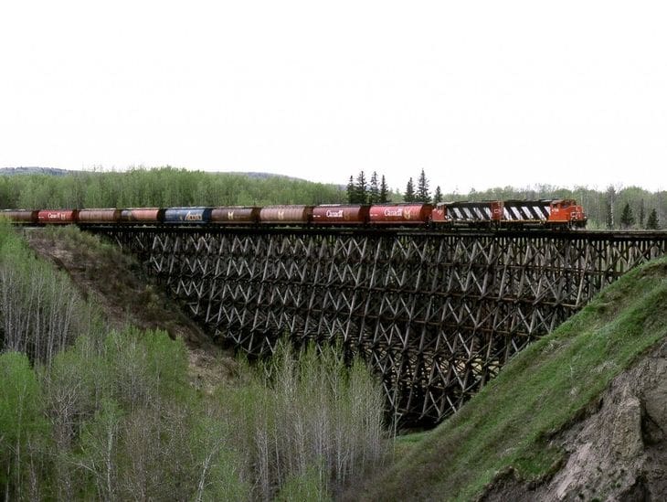 A long train crosses a tall wooden trestle bridge over a green valley.