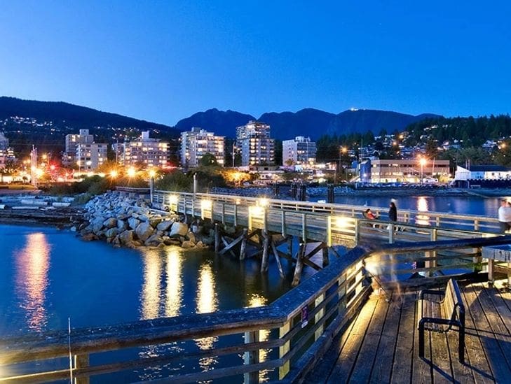 A city waterfront at dusk with illuminated buildings and a wooden pier.