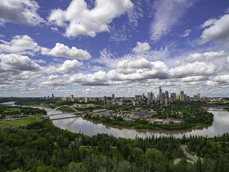 A city skyline with a river and lush greenery under a partly cloudy sky.