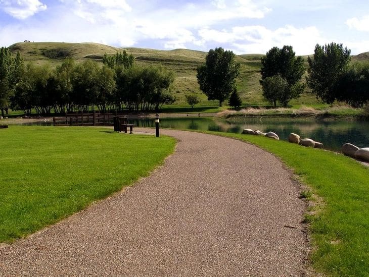 A winding gravel path curves through a green park with trees and a wooden bench.
