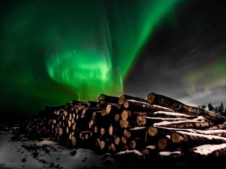Aurora borealis glowing above stacked logs in a snowy landscape at night.